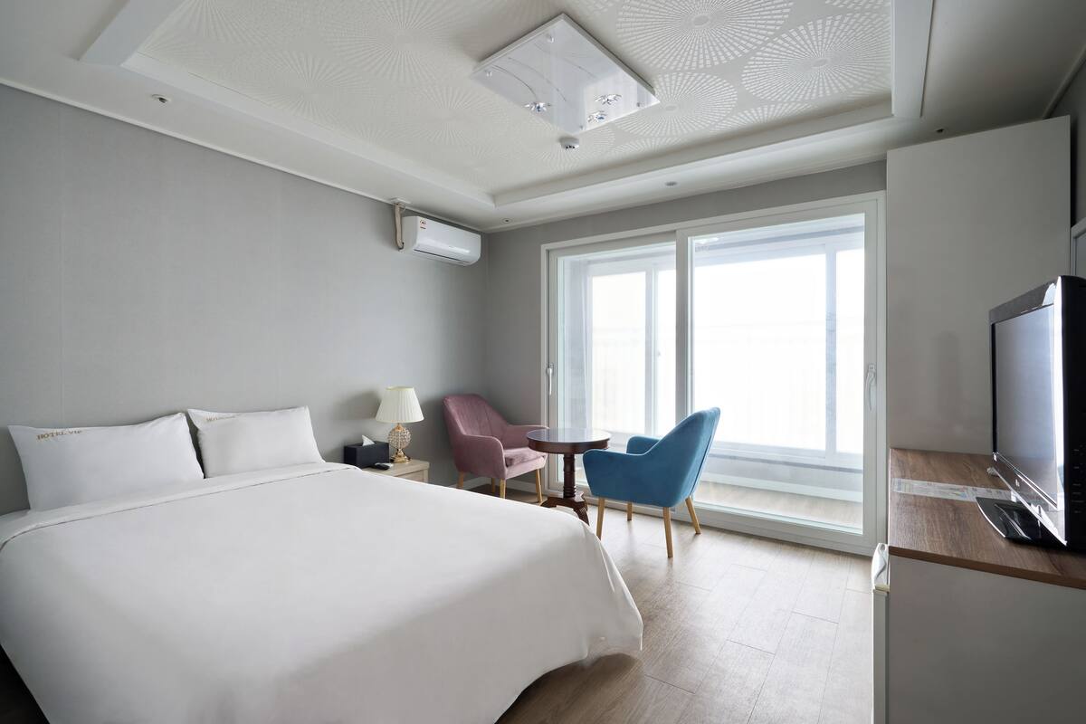 A hotel room with a large bed, a desk with a TV, a chair, and a window with a view.