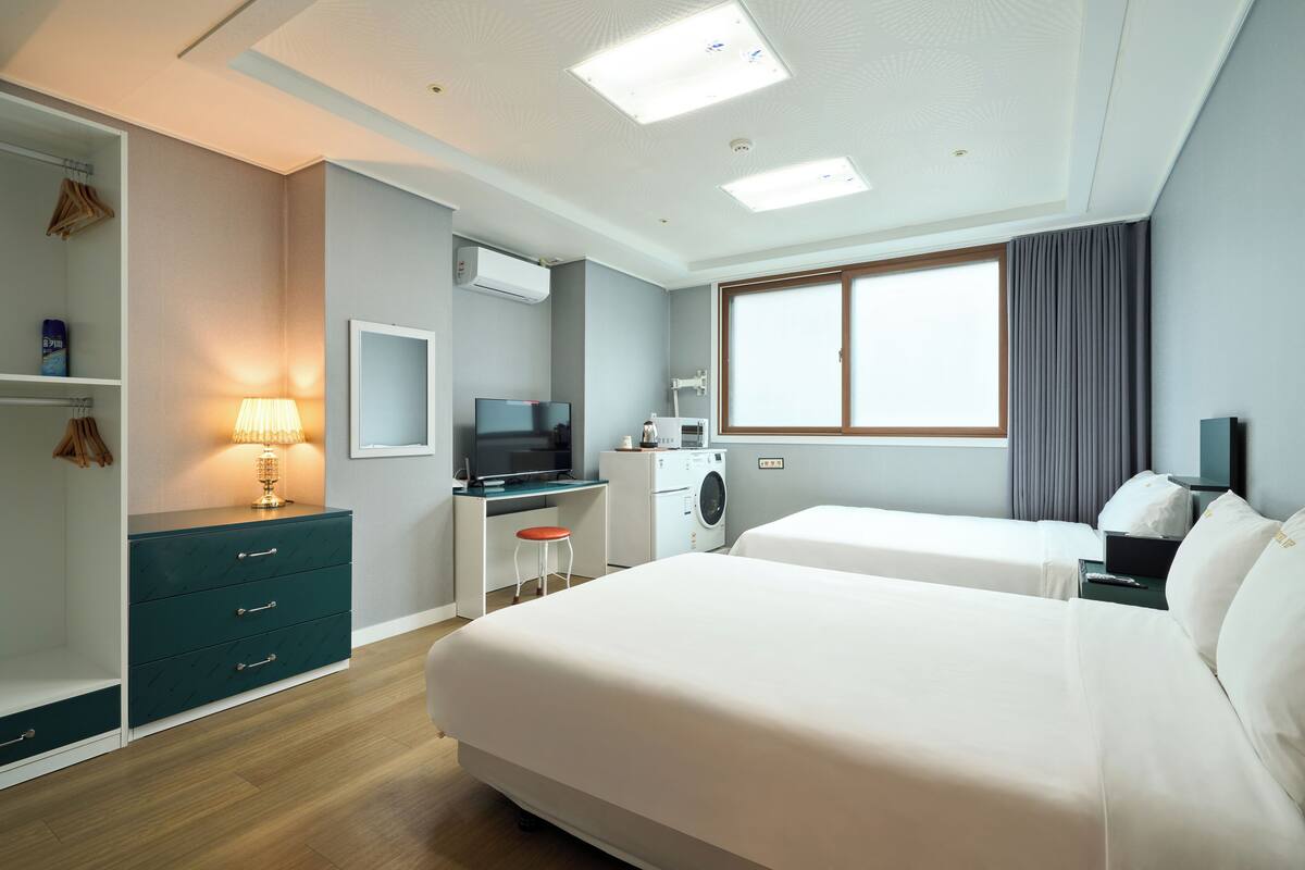 A hotel room with two beds, a desk with a computer, a washing machine, and a wardrobe.
