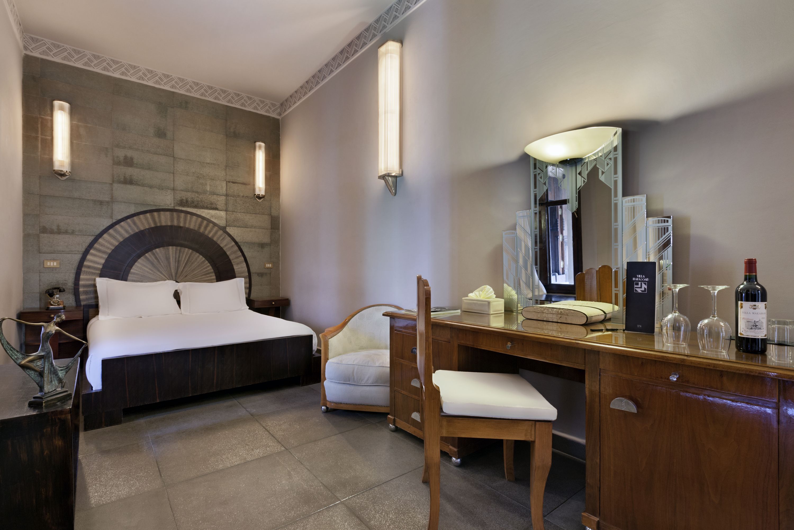 A hotel room with a bed, a desk, a chair, a dresser, a mirror, and a wine bottle.