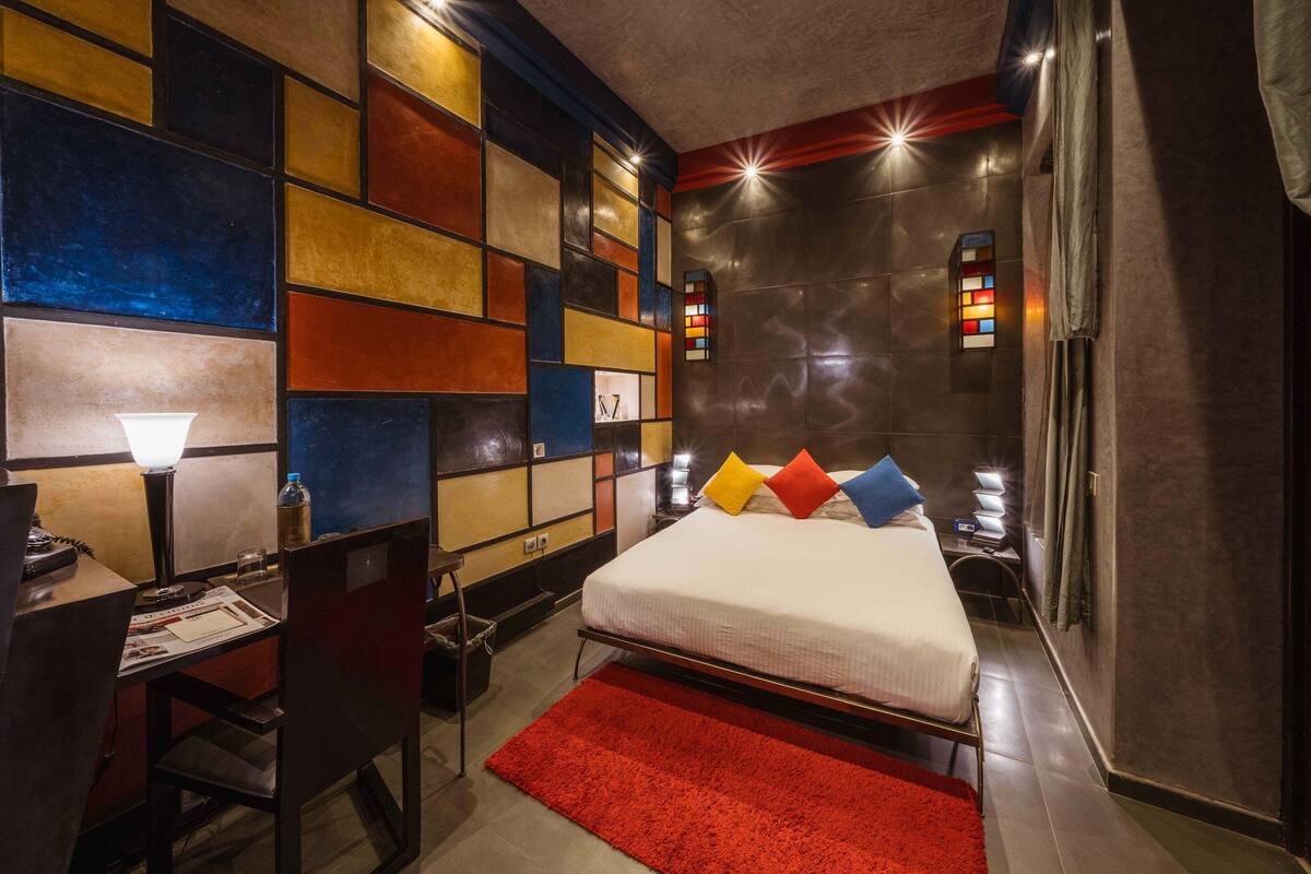 A modern hotel room with a large bed, a desk with a lamp, and a red rug.