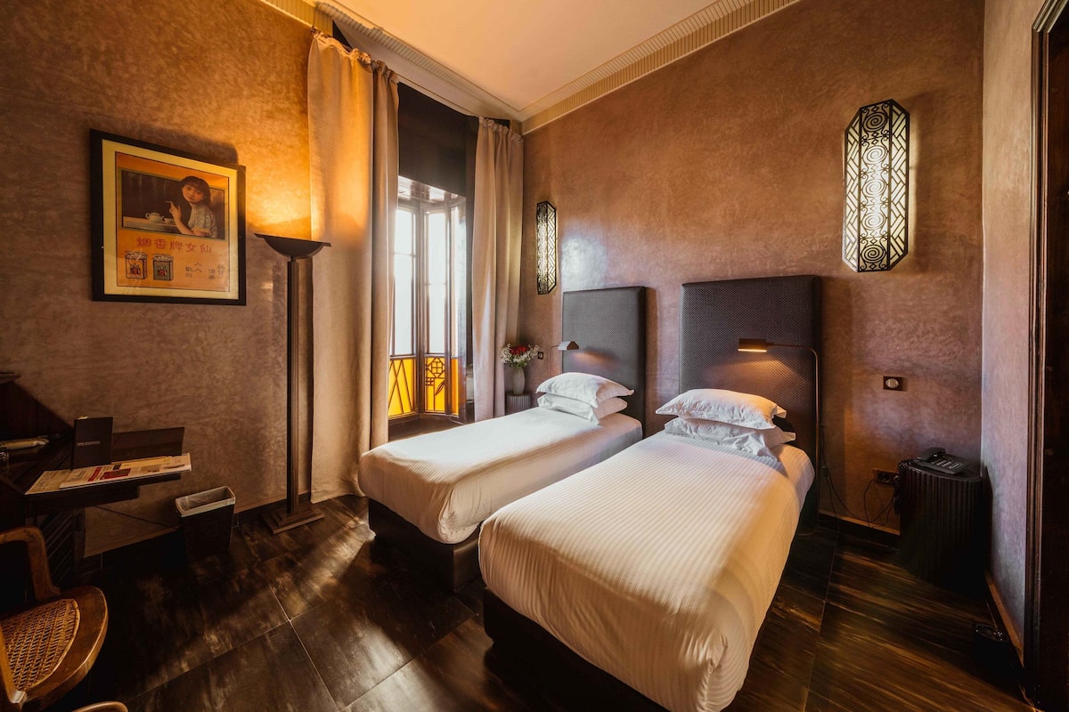 A hotel room with two beds, a desk with a chair, a lamp, and a framed picture on the wall.