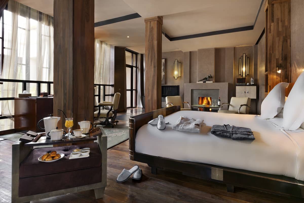 A modern hotel room with a large bed, a desk with breakfast items, a fireplace, and a view of the outdoors.
