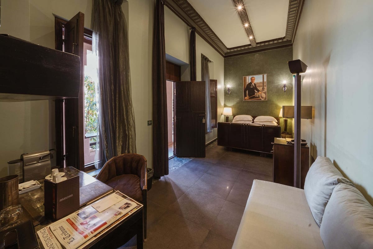 A hotel room with a bed, a desk, a chair, and a sofa. There is a painting on the wall and a window with curtains.