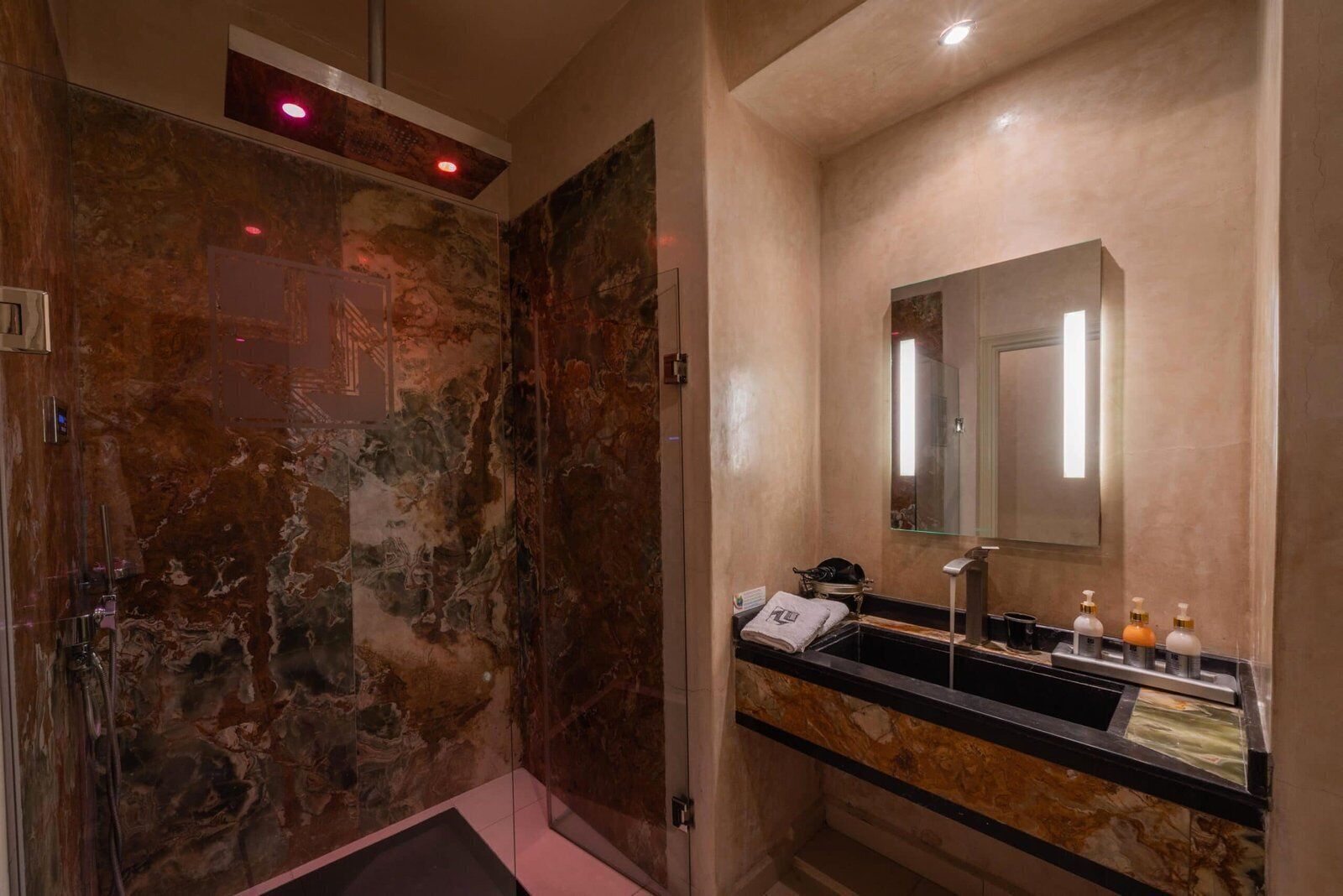Un baño moderno con pared de mármol, cabina de ducha de vidrio, lavabo con artículos de aseo y espejo.