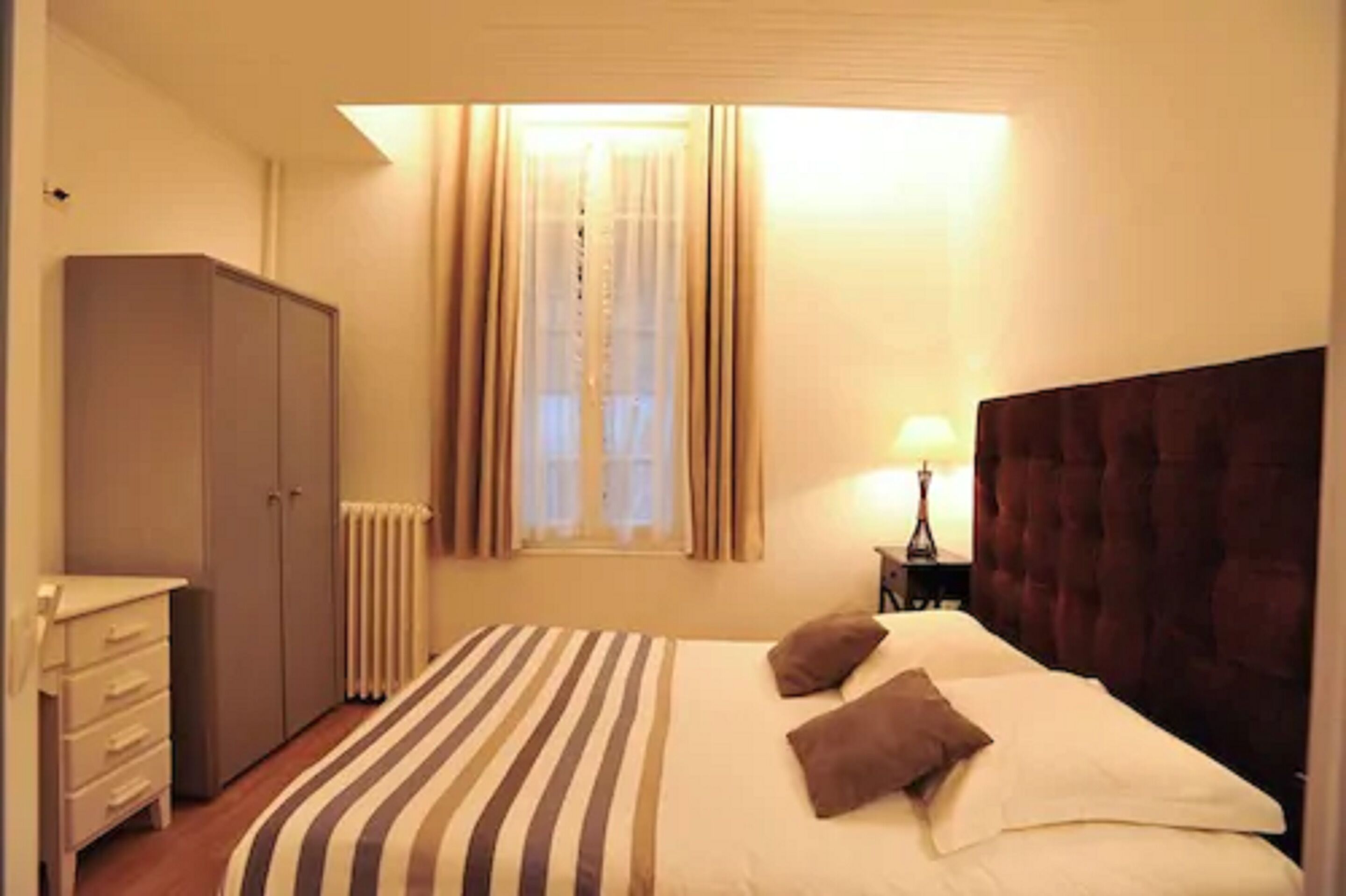 A bedroom with a bed, a wardrobe, a radiator, a window with curtains, and a nightstand with a lamp.