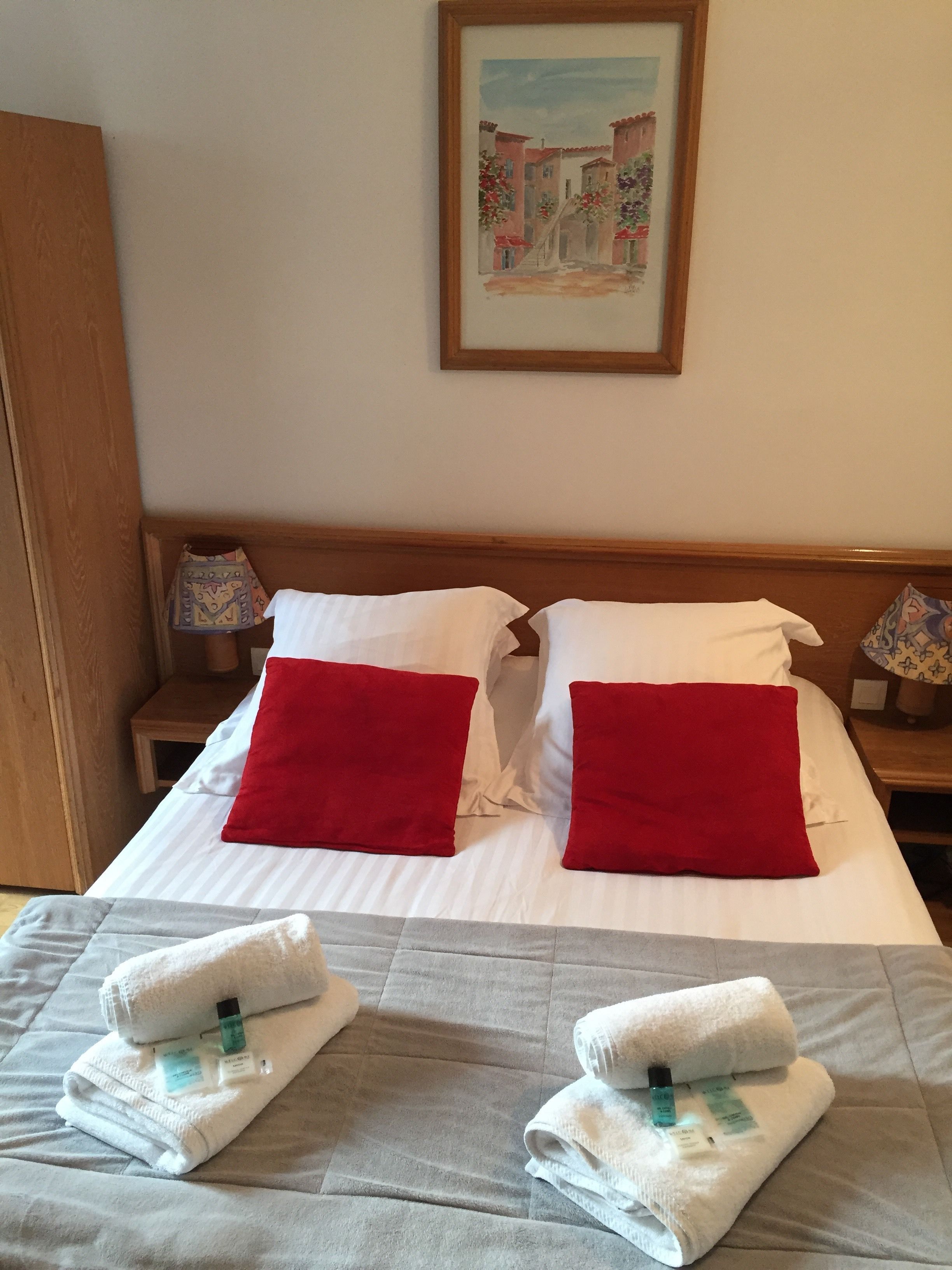 A neatly made bed with white and red pillows, a grey bedspread, and two folded towels with toiletries on a wooden headboard.