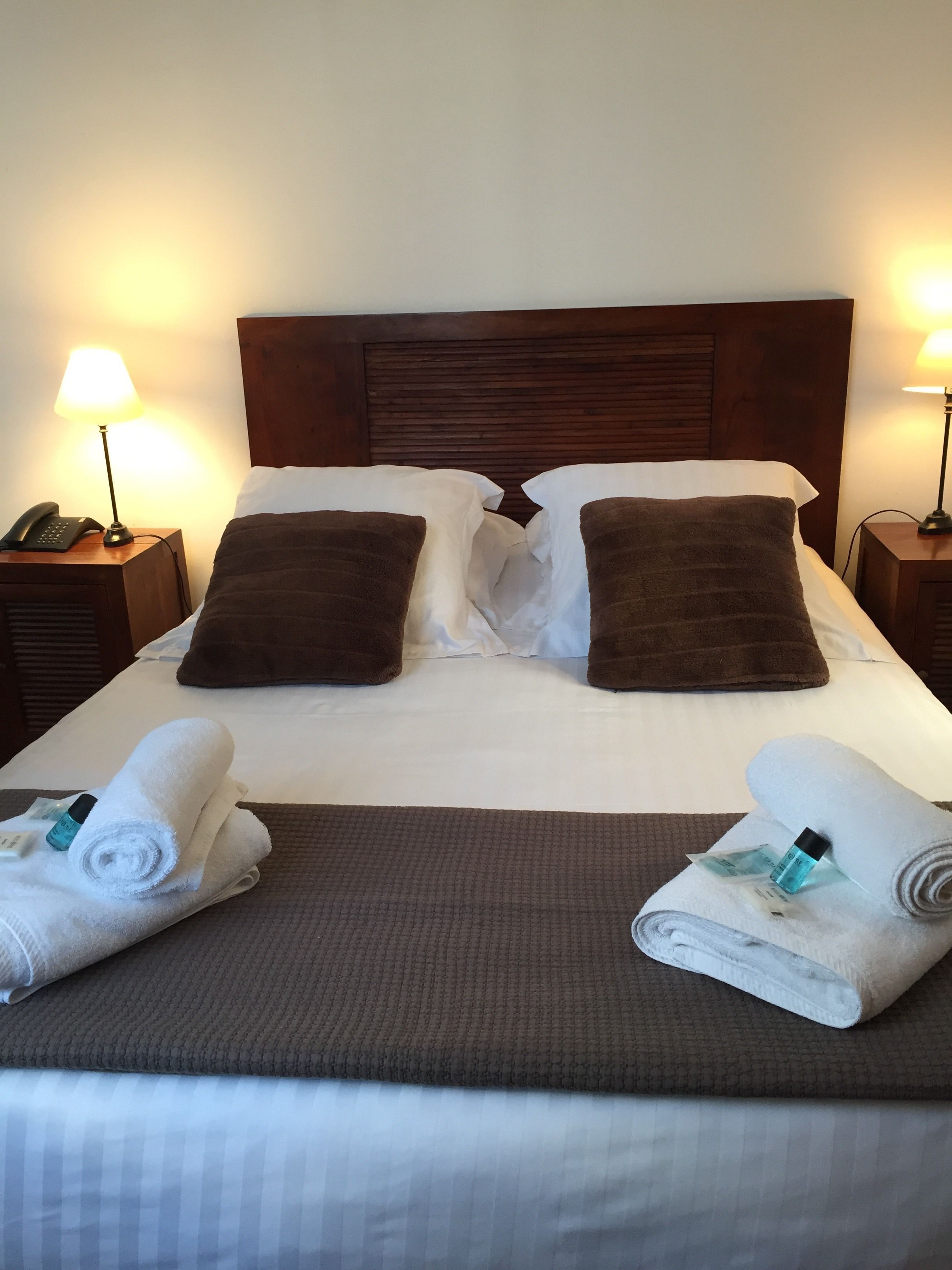 Una cama bien hecha con un cabecero marrón, dos almohadas y dos toallas enrolladas a un lado.