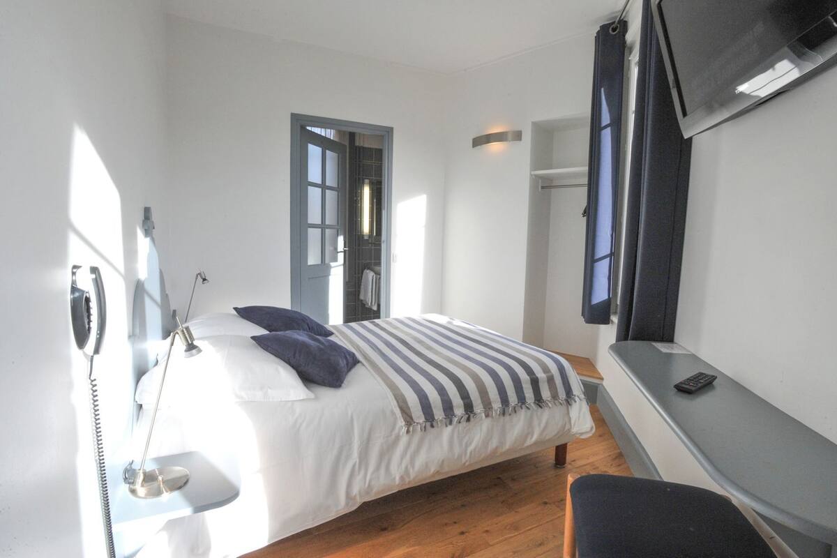 A bedroom with a bed, a desk, a chair, a TV, a telephone, and a window.