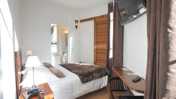 A hotel room with a bed, bedside table with a lamp and phone, a desk with a chair, a TV mounted on the wall, and a wooden door.