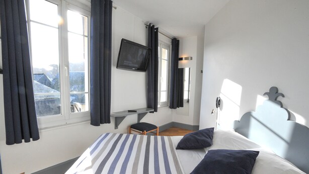 A hotel room with a bed, a desk, a chair, a TV, and a window with curtains. - Hôtel des Falaises (Etretat)