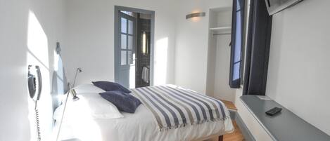 A bedroom with a bed, bedside table, and a desk with a chair. There is a telephone on the wall and a window with curtains.