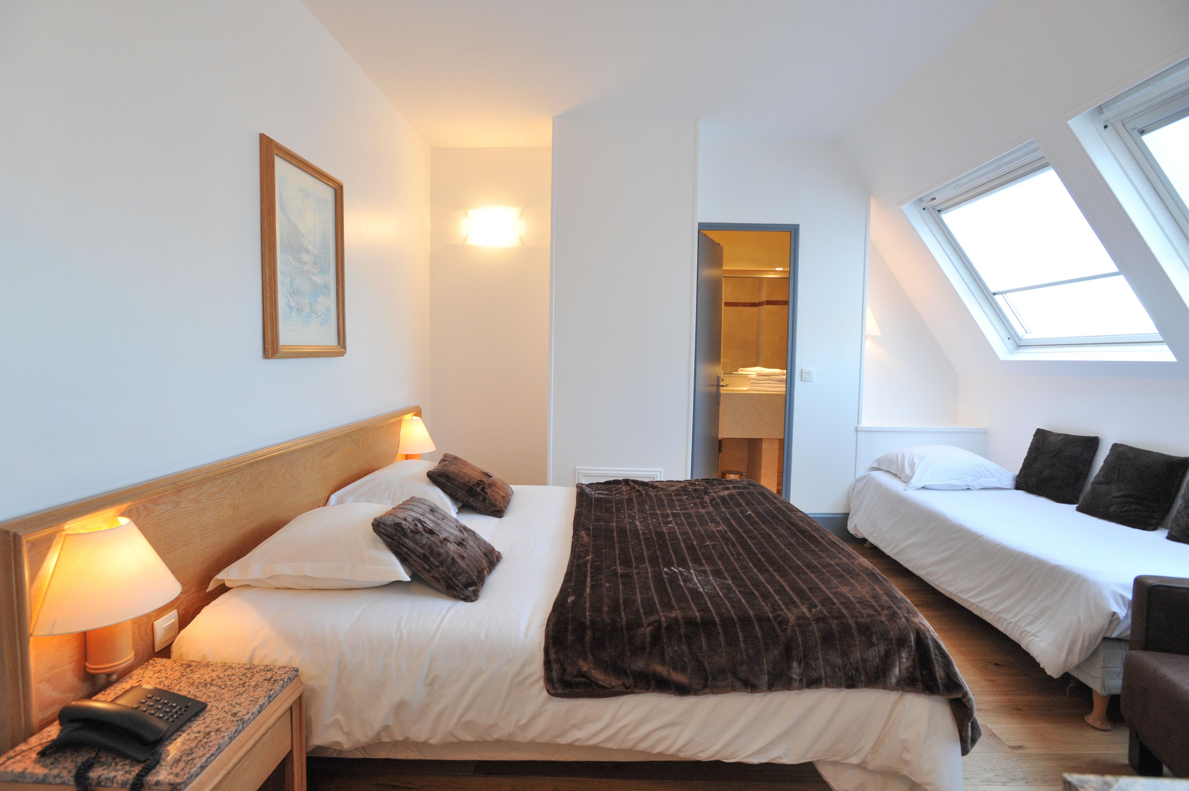 A hotel room with a large bed, a smaller bed, a desk with a lamp, a telephone, and a skylight.