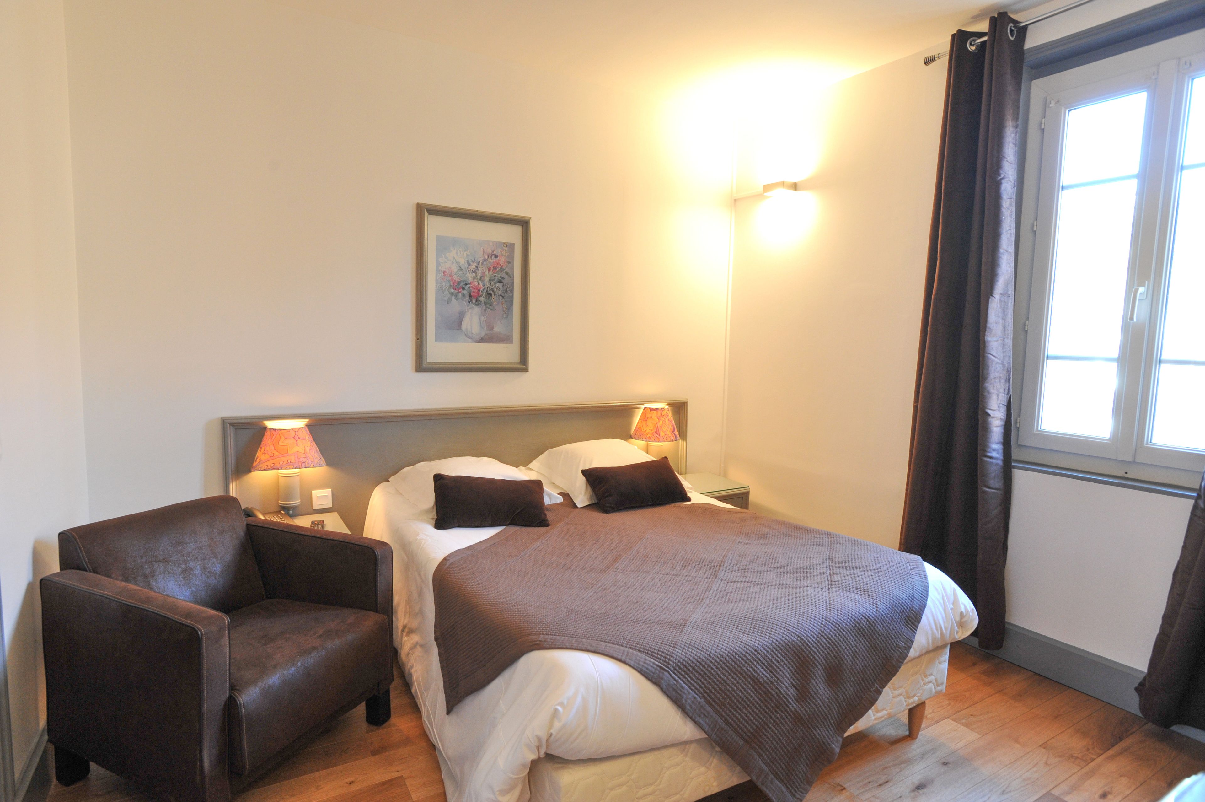 A hotel room with a bed, two bedside lamps, a chair, a framed picture on the wall, and a window with curtains.