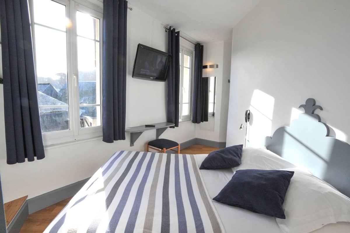 A hotel room with a bed, a desk, a chair, a television, and a view of buildings outside the window.