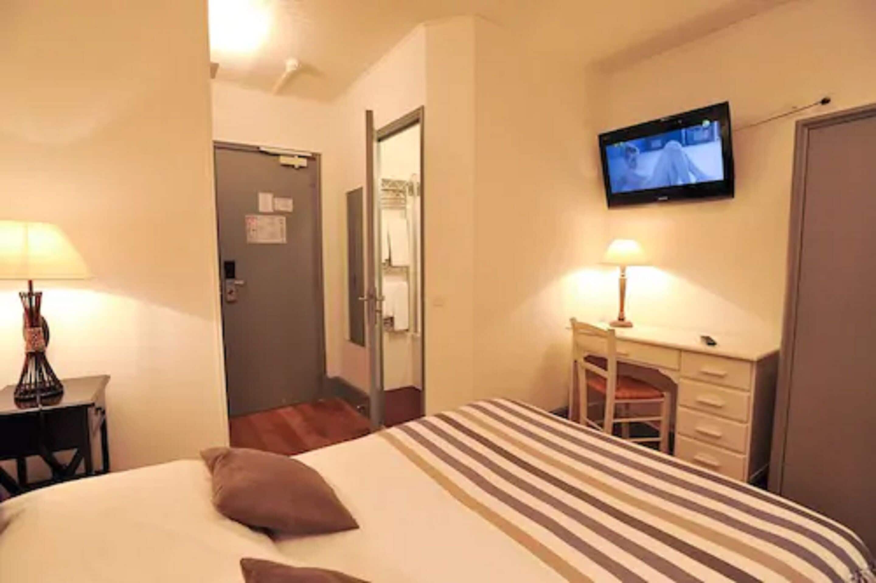 Habitación de hotel con cama, dos lámparas de noche, escritorio con silla, televisor montado en la pared y puerta que da a otra habitación.