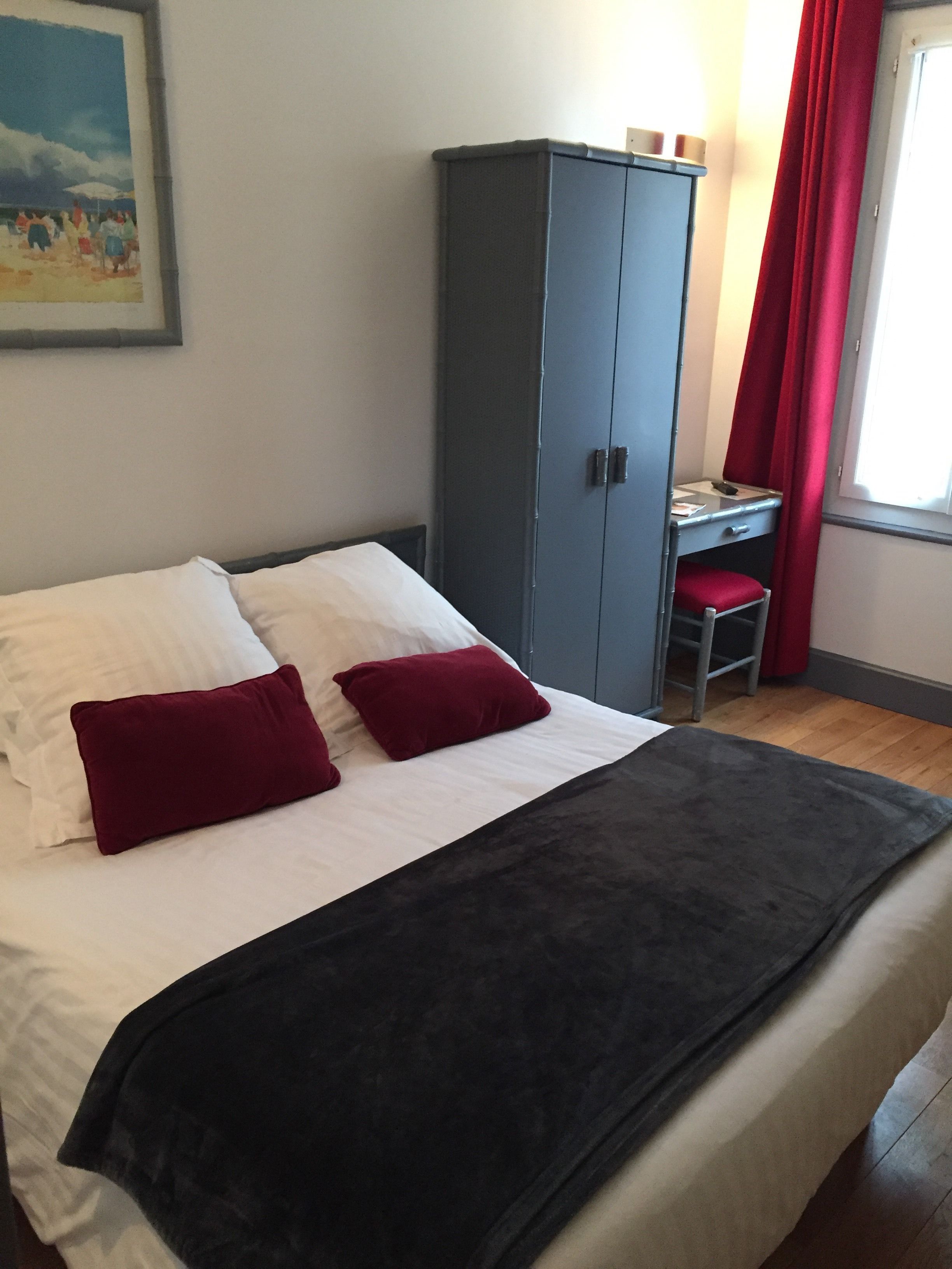 Een bed met wit en rood beddengoed, een grijze garderobe, een bureau met stoel en een raam met rode gordijnen.