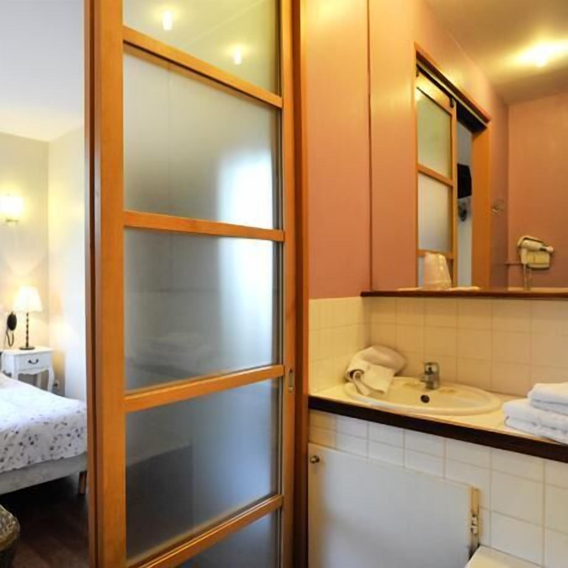 A hotel room with a bed, a bedside table with a lamp, a bathroom with a sink, a mirror, and a towel rack.