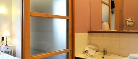 Una habitación de hotel con una cama, una mesita de noche con lámpara, un baño con lavamanos, un espejo y un tendedero para toallas.