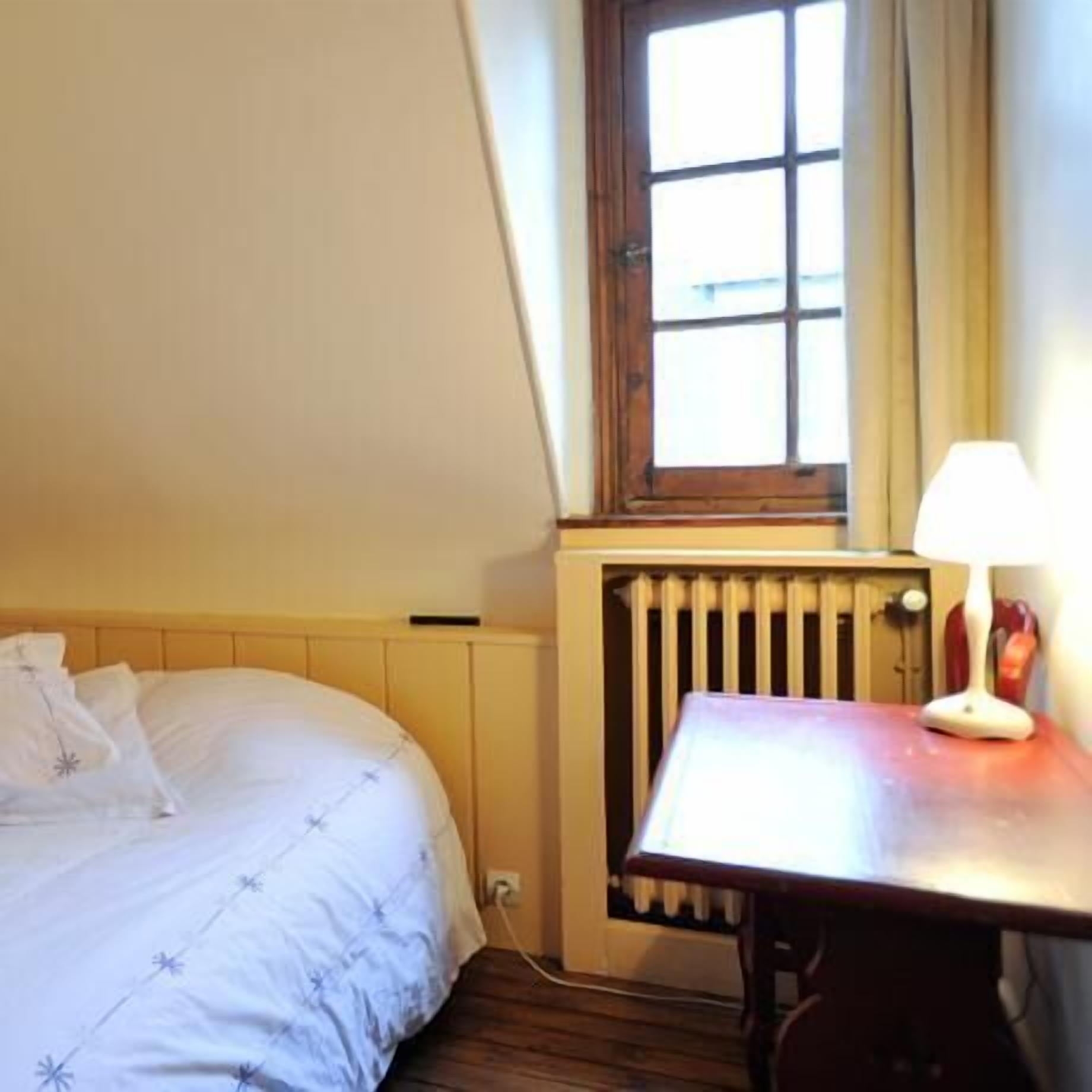 A bedroom with a bed, a table with a lamp, a window, and a radiator.