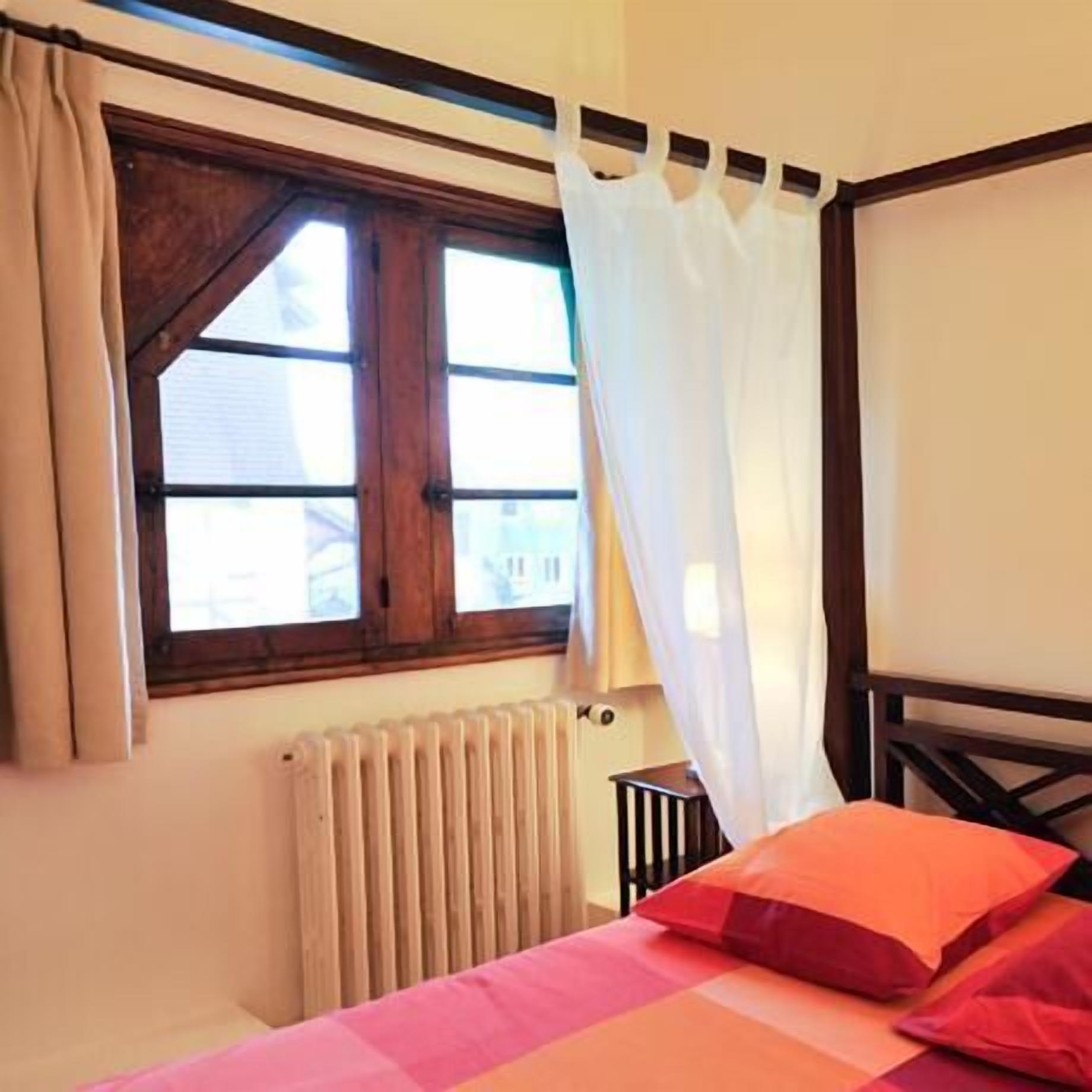 A bedroom with a bed, a window with curtains, a radiator, and a wooden headboard.