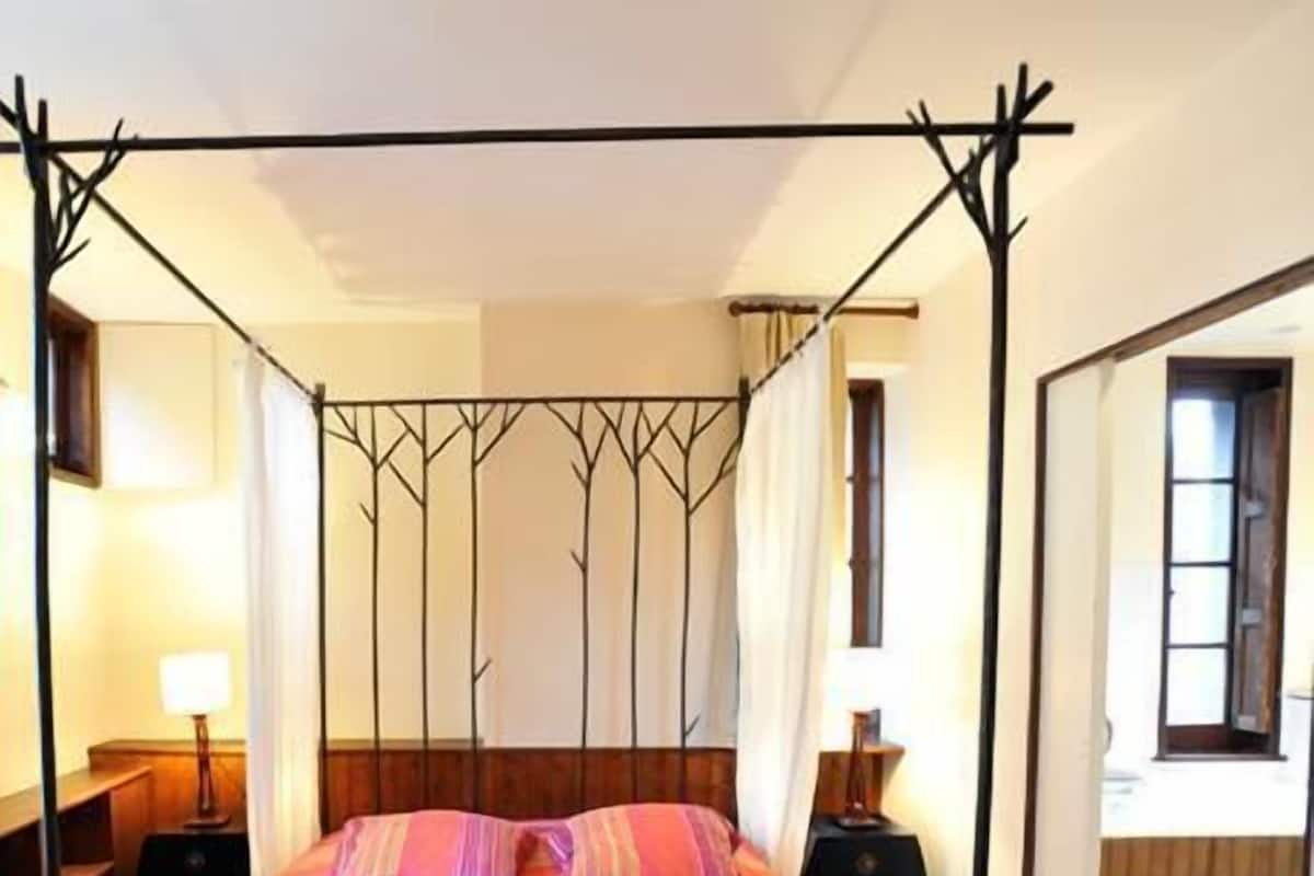 A four-poster bed with a striped bedspread, two bedside tables with lamps, and a view of the room's interior through a doorway.