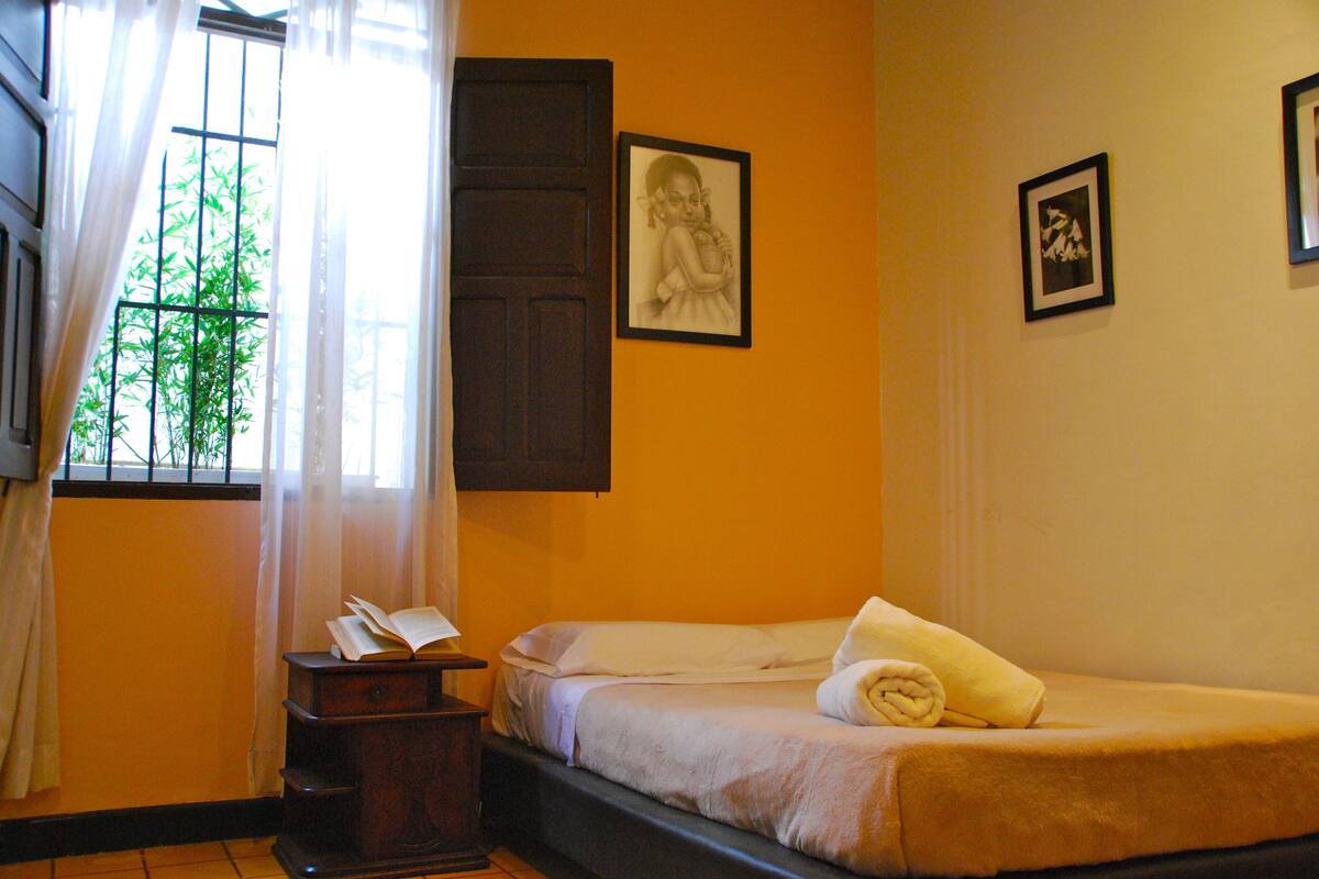 Una habitación con dos camas, una ventana con cortinas, un escritorio con un libro y cuadros en la pared.