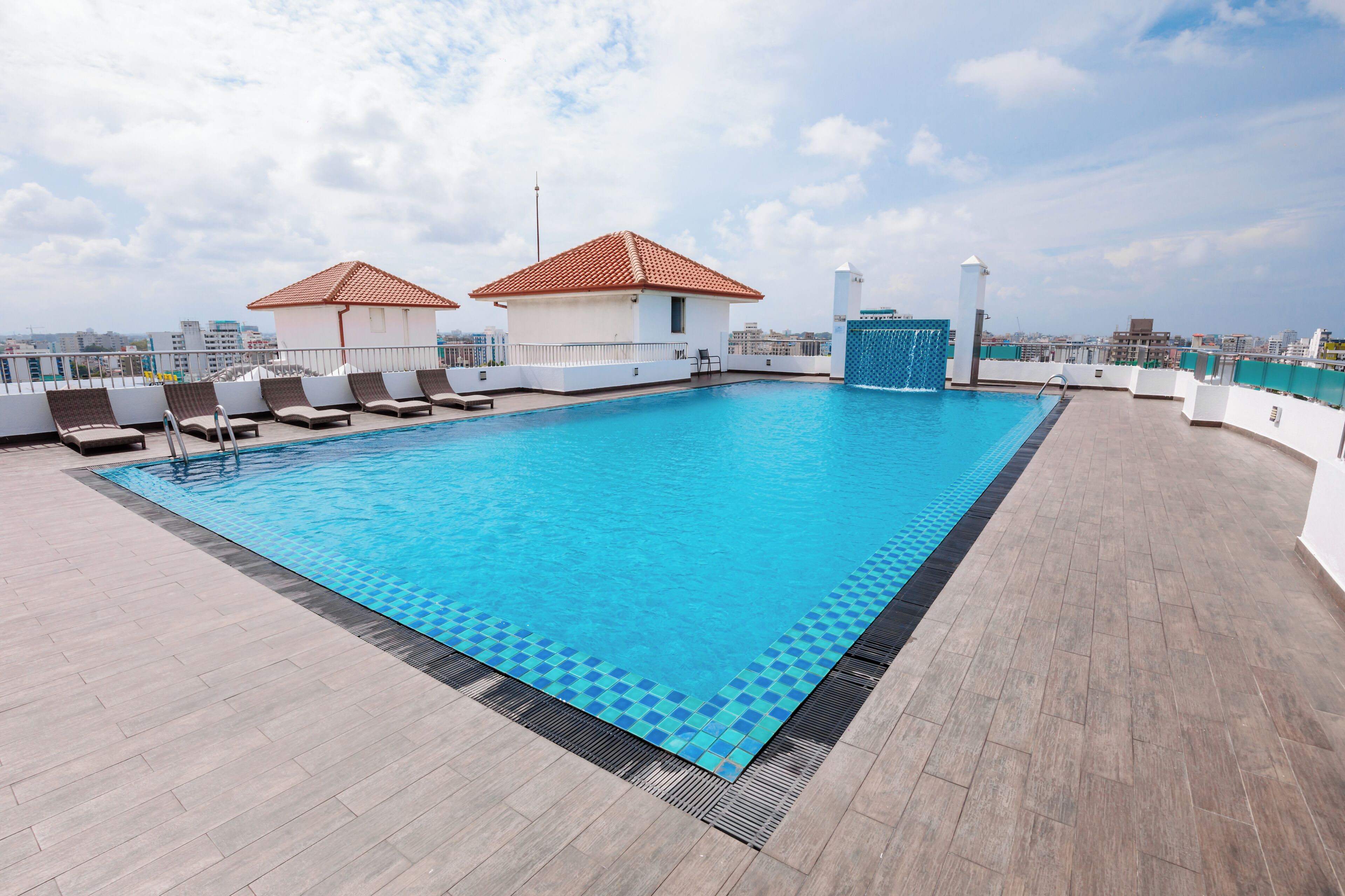 A rooftop pool with lounge chairs, a small building with a red roof, and a city view.