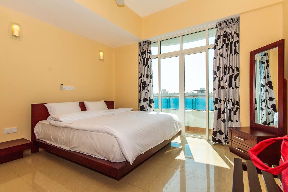 Une chambre d’hôtel avec un grand lit, une table de chevet en bois, une chaise rouge et une vue sur la mer depuis le balcon.