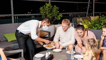 Un gruppo di persone cena all'aperto di notte, con un tavolo apparecchiato per un pasto, circondato da vegetazione e un corrimano.