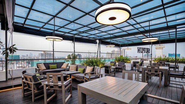 An outdoor seating area with wooden furniture and a glass ceiling, overlooking a cityscape. - Supun Arcade Residency (Colombo)