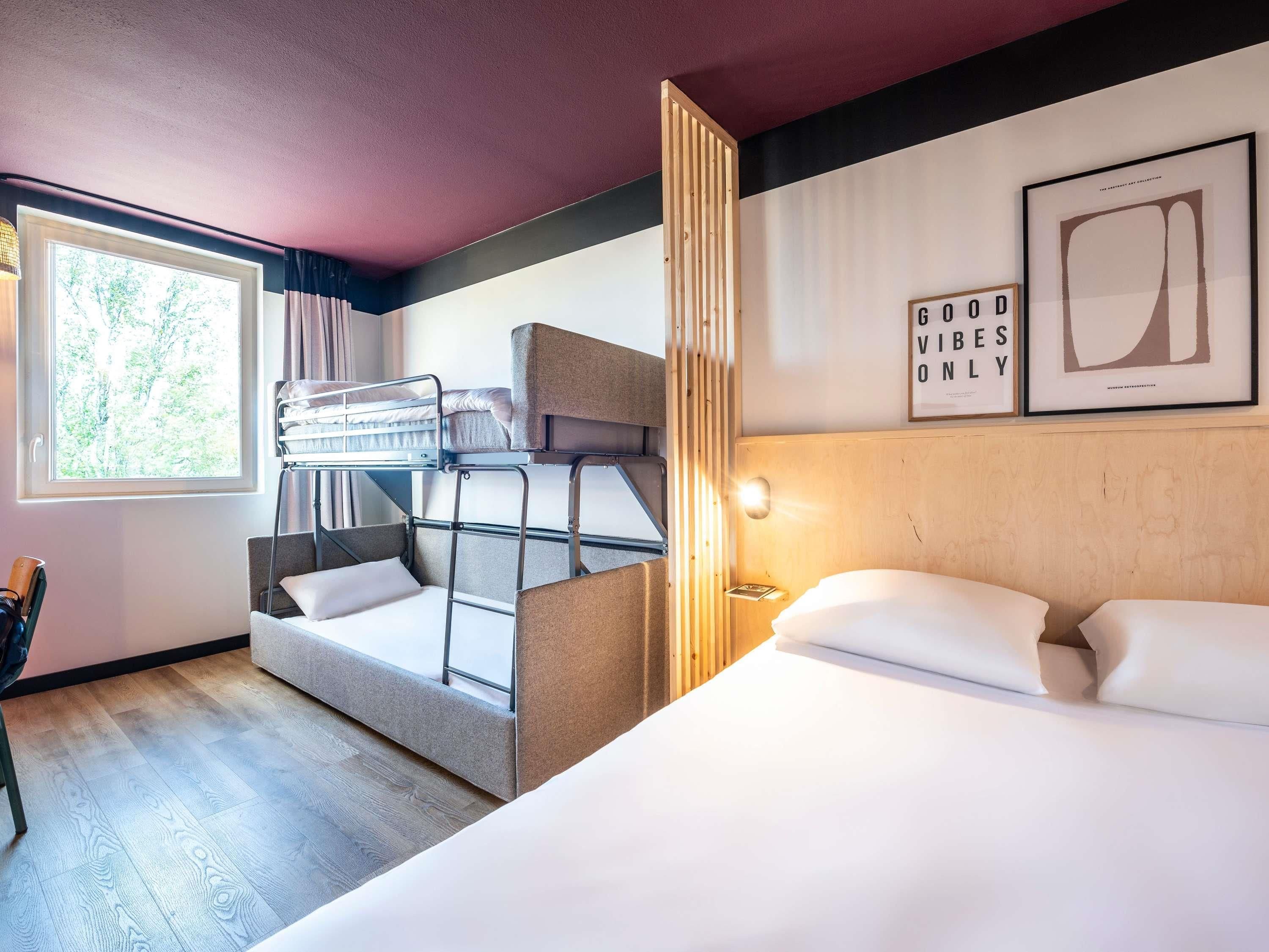 Une chambre avec des lits superposés, un grand lit, une fenêtre donnant sur la forêt et un mur orné de tableaux.