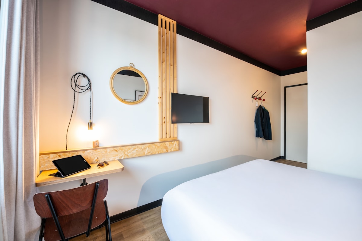 A hotel room with a bed, a desk with a tablet and sunglasses, a mirror, and a TV mounted on the wall.