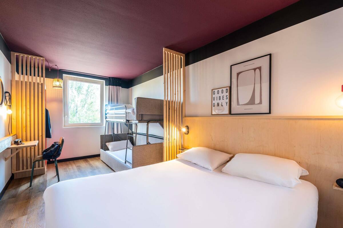 A hotel room with a bunk bed, a desk with a chair, a window with curtains, and a wall with framed artwork.