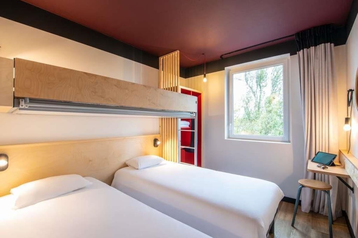 A hotel room with two beds, a desk, a window with curtains, and a red cabinet.