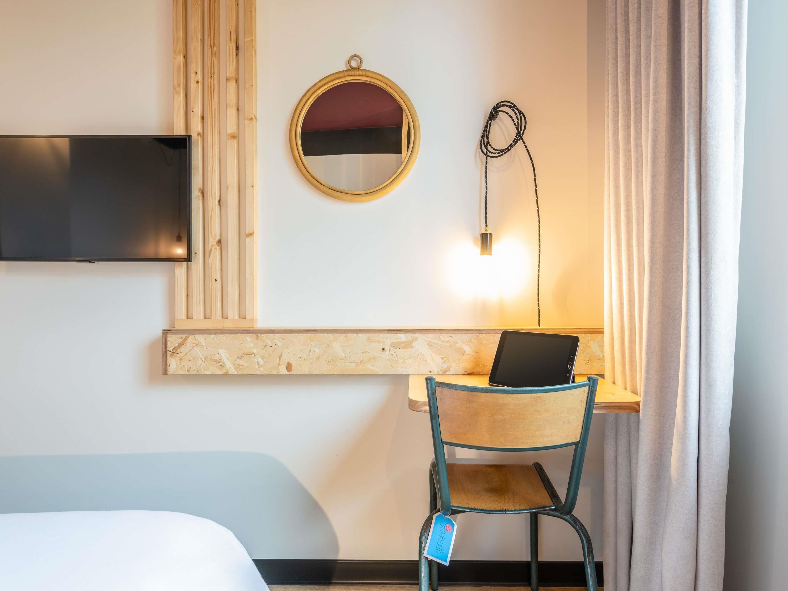 Une chambre d’hôtel équipée d’un lit, d’un bureau avec un ordinateur portable, d’une télévision, d’un miroir et d’une lampe fixée au mur.