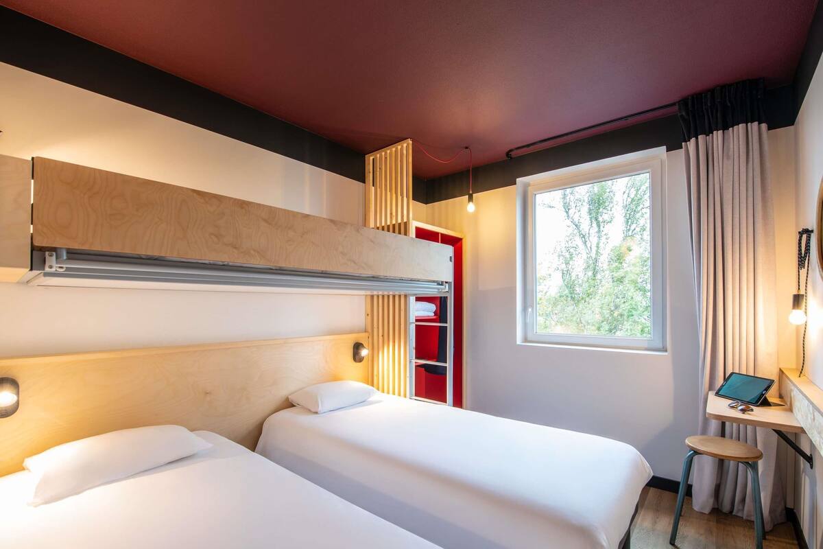 A hotel room with two beds, a desk with a chair, a window with curtains, and a red storage unit.