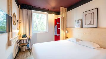 A hotel room with a bed, a desk with a chair, a TV, a window with curtains, and a red wardrobe.
