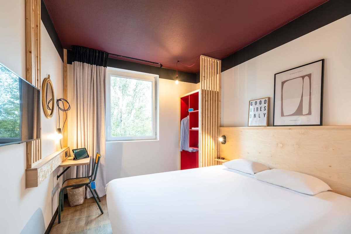 A hotel room with a bed, a desk with a chair, a TV, a window with curtains, and a red wardrobe.