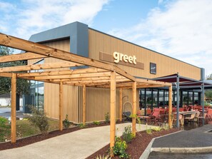Un edificio moderno con un letrero que dice 'greet', una pérgola de madera, asientos exteriores y una entrada de vidrio.