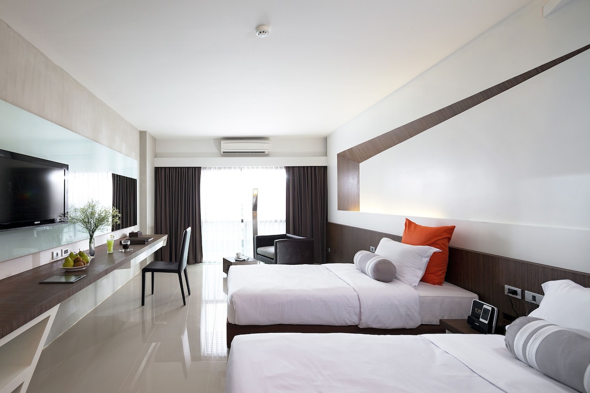 A hotel room with a flat-screen TV, a dining area, two beds with pillows, a desk with a laptop, and a window with curtains.