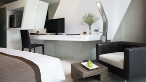 Moderni hotellihuone, jossa on sänky, työpöytä tietokoneella, tuoli, sohvapöytä ja huonekasvi.