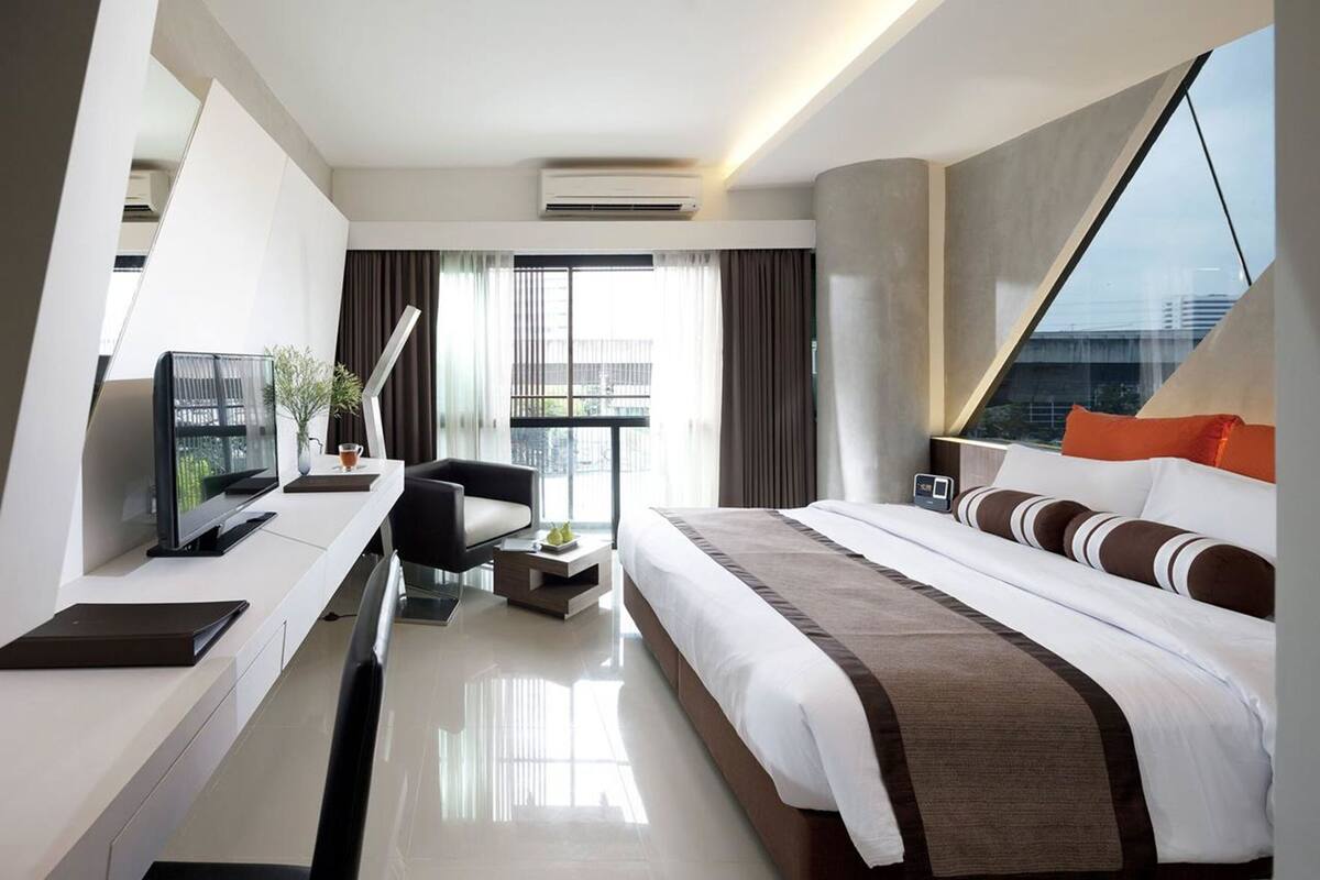 A modern hotel room with a large bed, a desk with a flat-screen TV, a chair, and a small table with a plant.