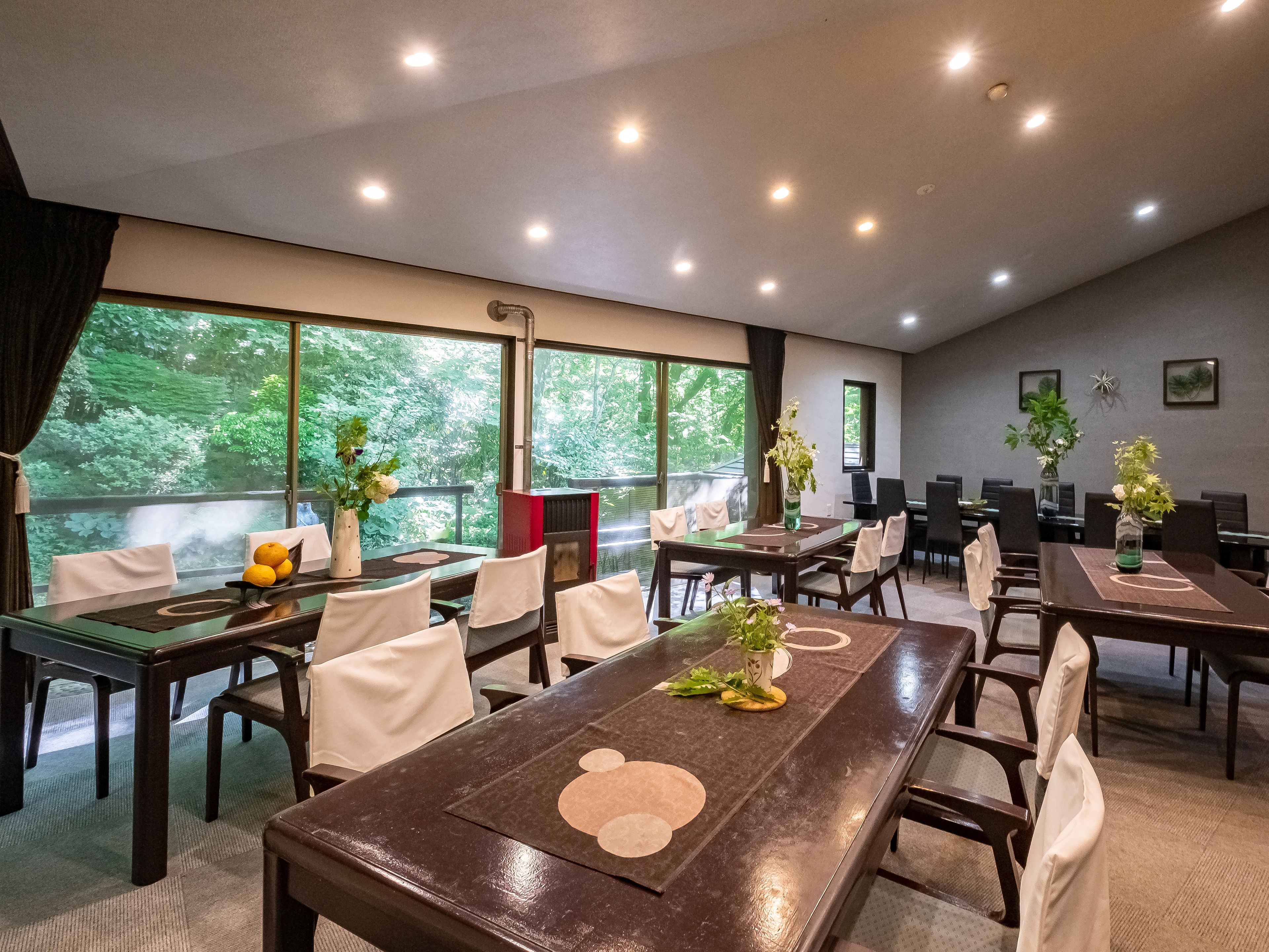 A conference room with multiple tables and chairs, large windows with a view of greenery, and ceiling lights.