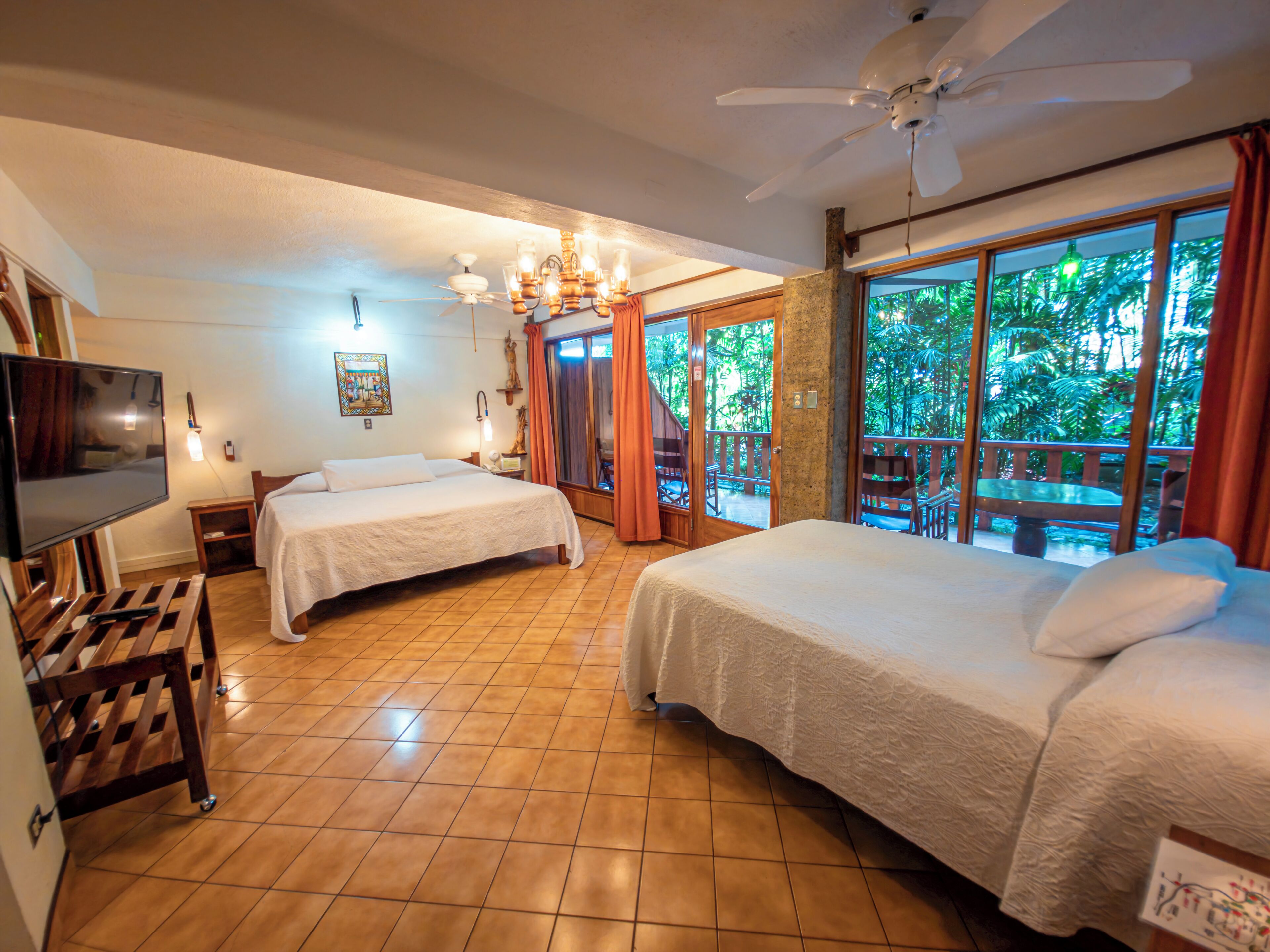 A hotel room with two beds, a television, a ceiling fan, and a balcony with a view of the outdoors.