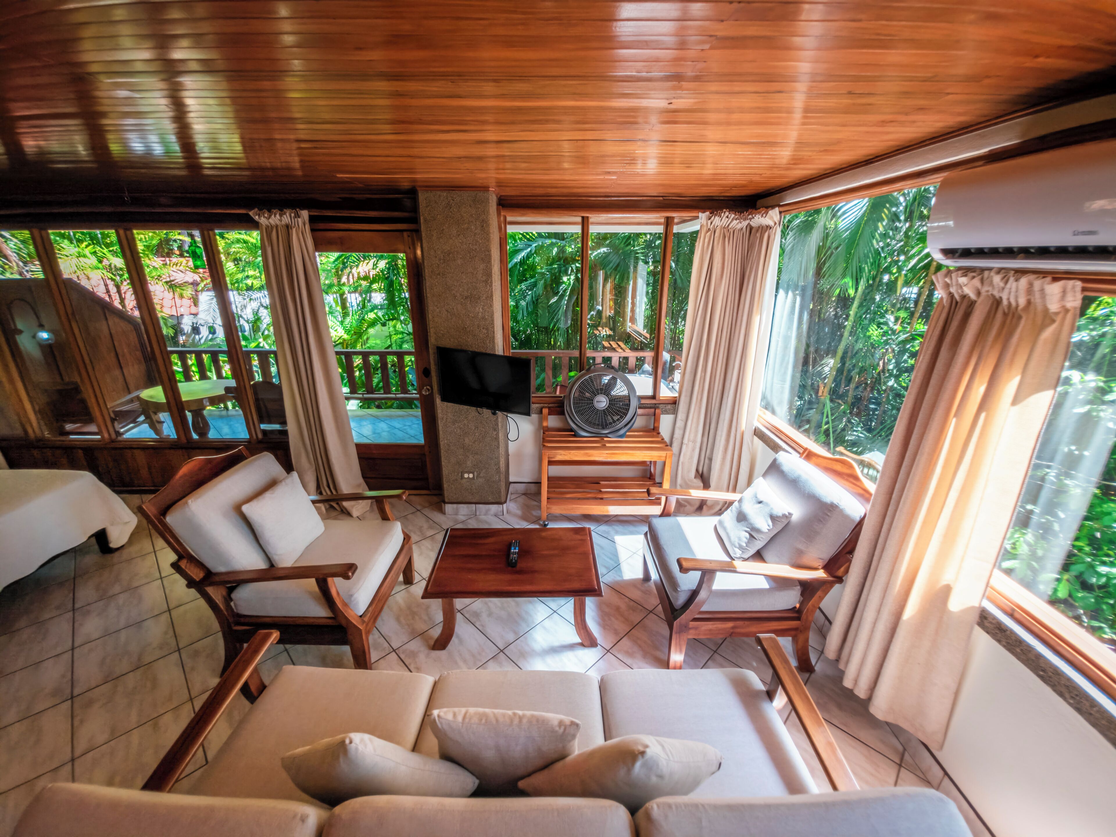 A room with a wooden ceiling, a sofa, two chairs, a TV, a fan, and large windows with curtains.
