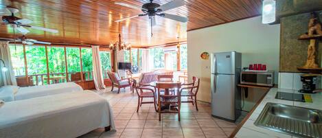 Una habitación con cama, una mesa de comedor con sillas, una zona de cocina con refrigerador y microondas, y un ventilador de techo.