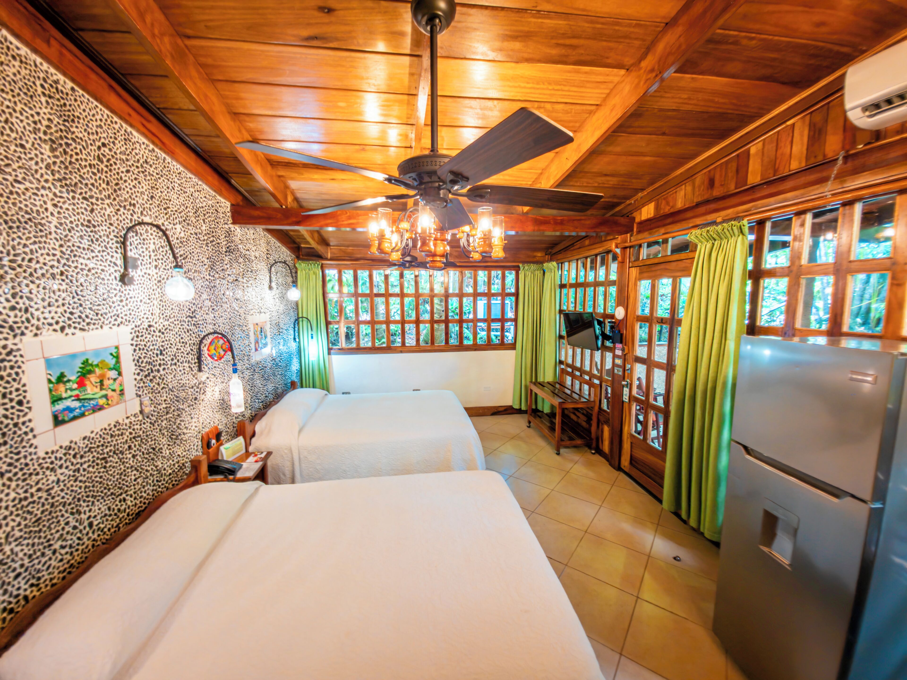 A room with two beds, a ceiling fan, a refrigerator, and a window with curtains.