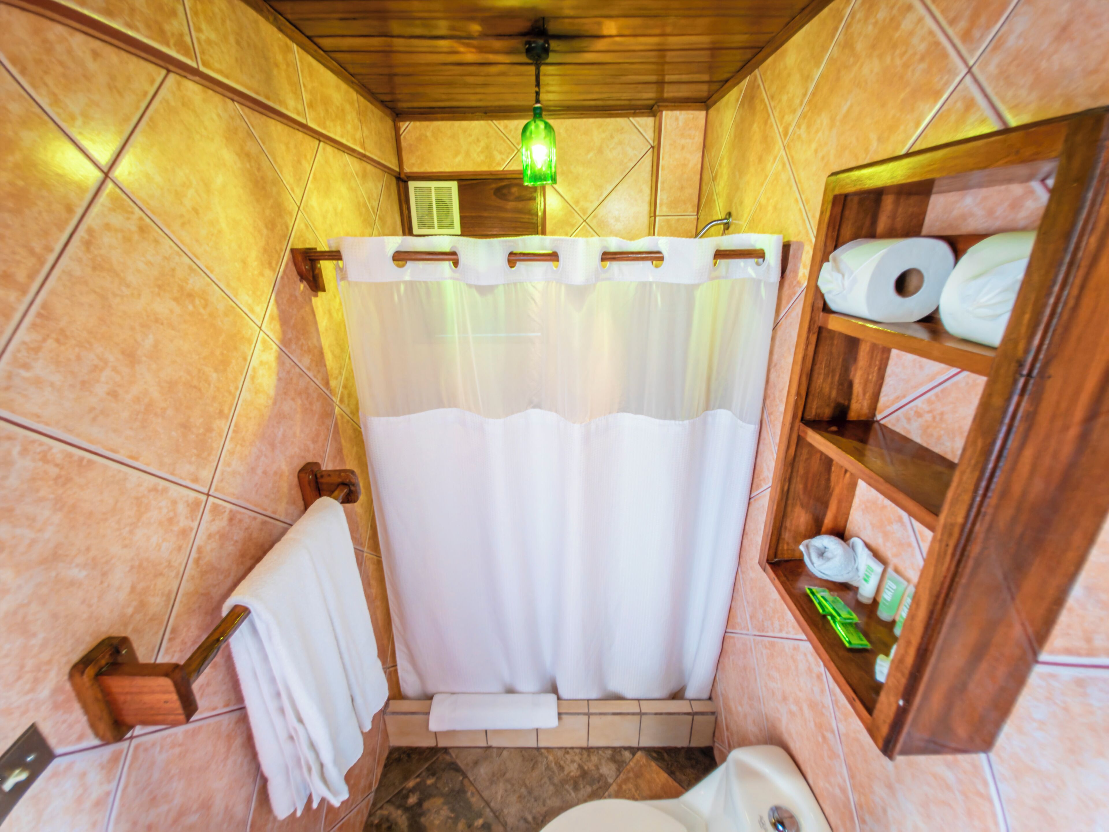 Une salle de bain avec un plancher en bois, des toilettes blanches, une étagère en bois contenant des articles de toilette et un rideau de douche.