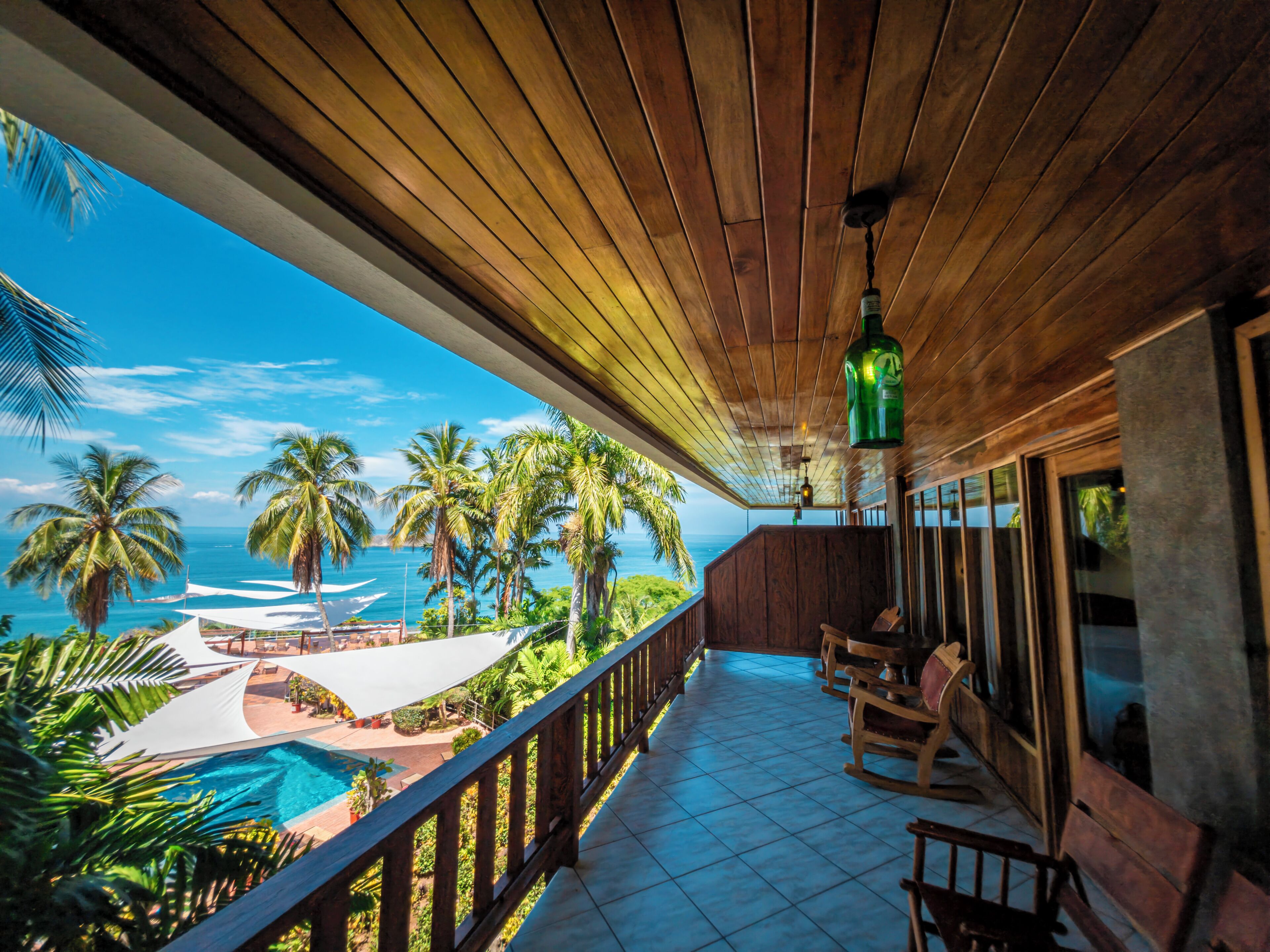 A wooden balcony with a view of the ocean, a pool, and palm trees.