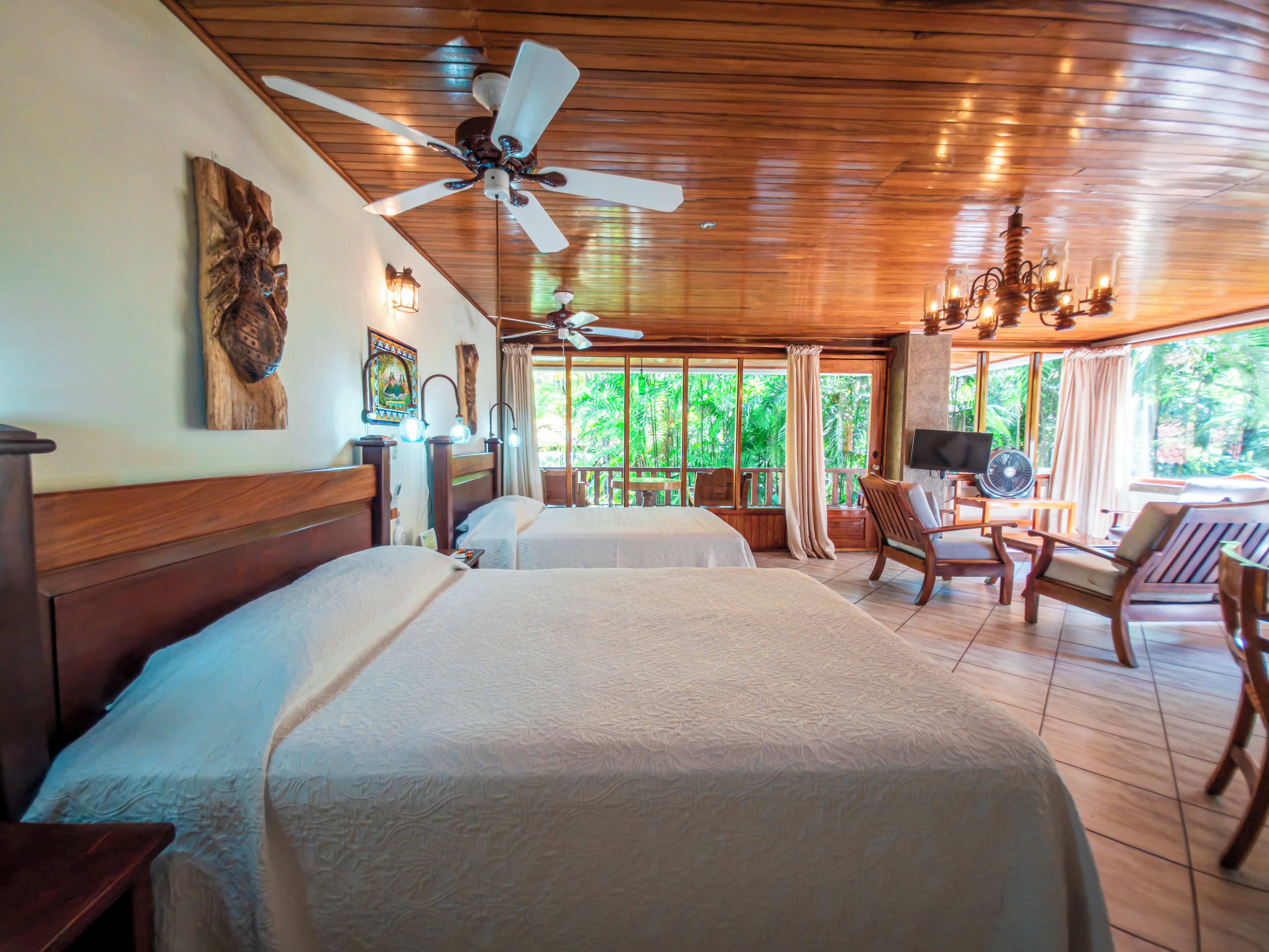 A hotel room with two beds, ceiling fans, a TV, and a balcony with a view of greenery.